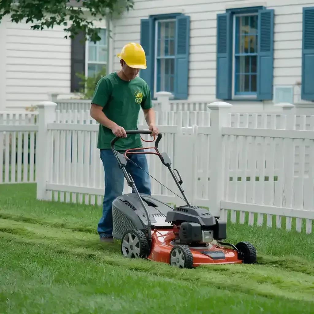 Worker mowing lawn with a red push mower beside a white fence and blue-shuttered house