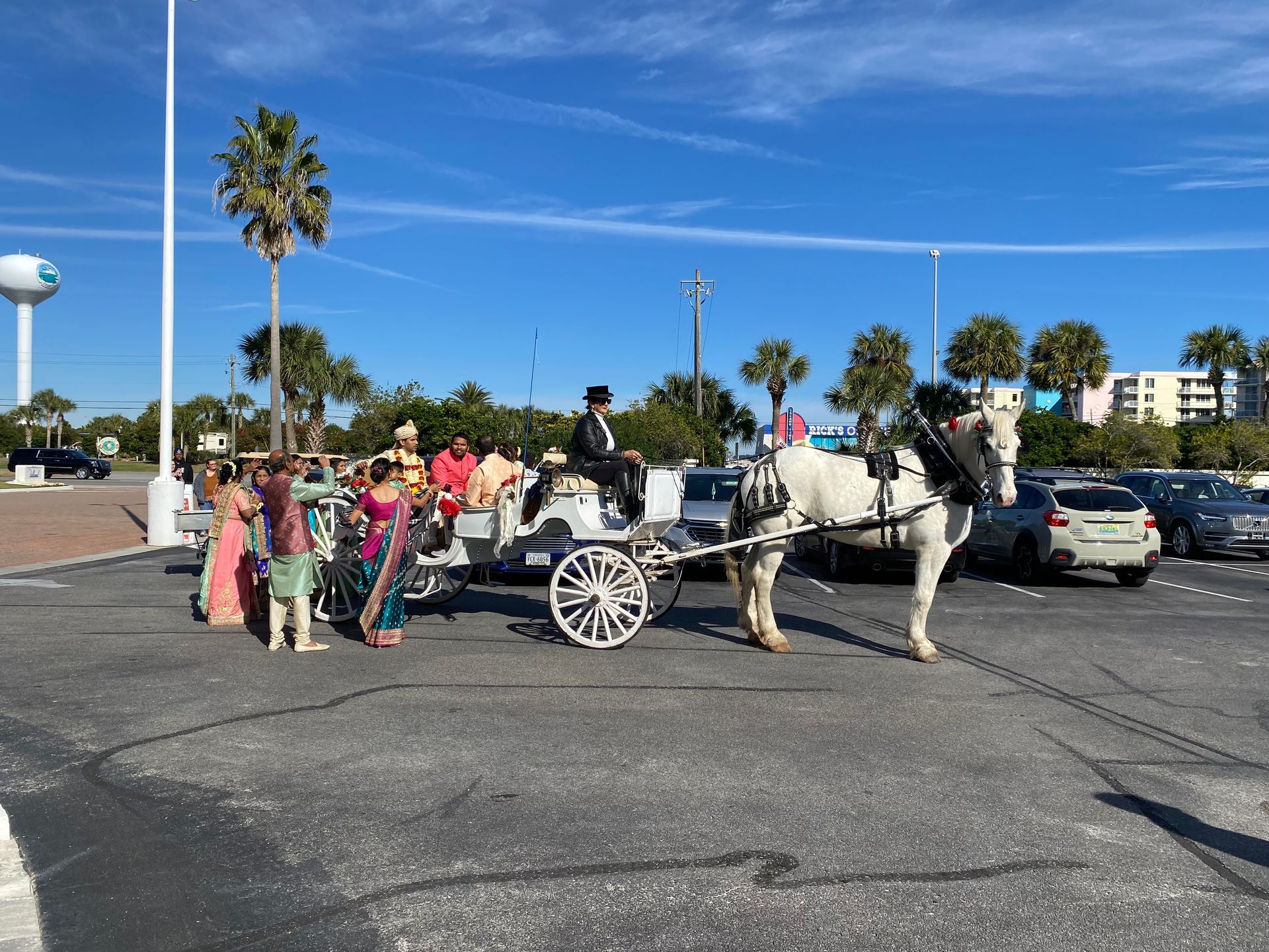 Horse-drawn carriage with passengers and pedestrians in a sunny parking lot near palm trees.
