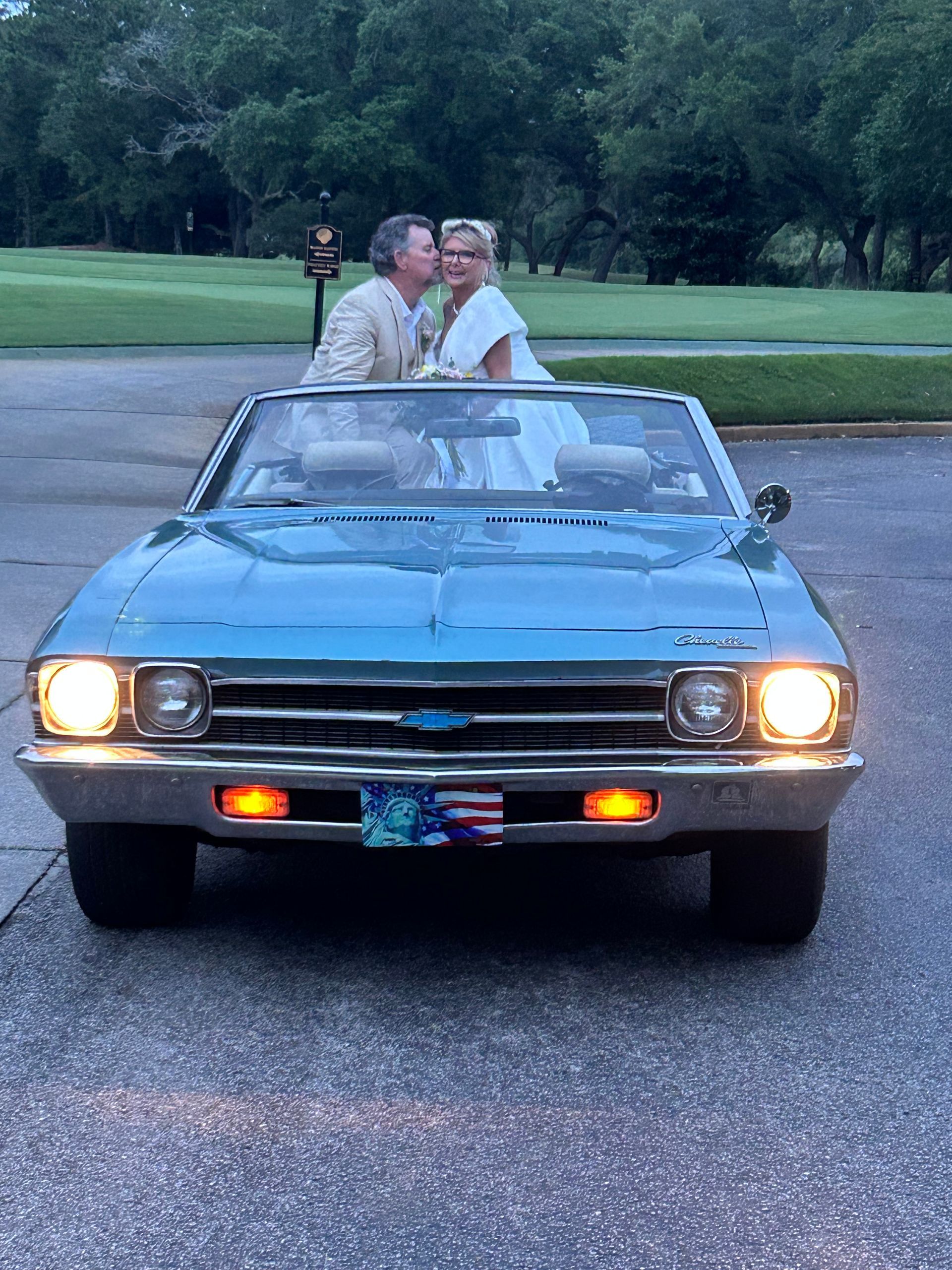 Couple standing in a classic convertible at dusk on a quiet paved road, headlights on.