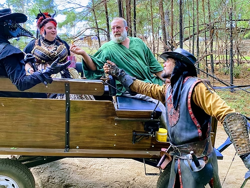Three people in colorful medieval costumes on a wooden wagon in a wooded outdoor setting