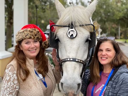 Two women smiling beside a white horse with festive red and fur-trimmed hats