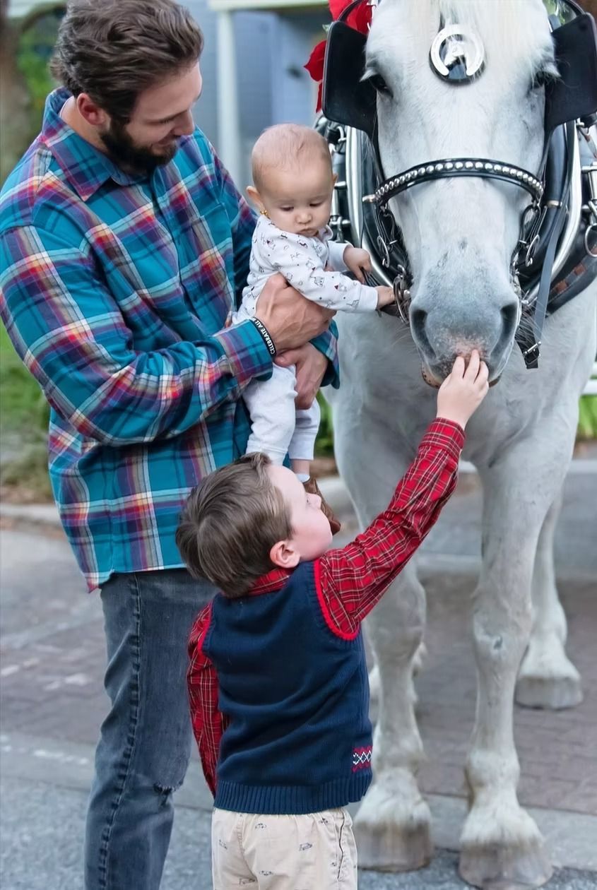 Man in plaid shirt holding a baby as a child pets a white horse on a city street