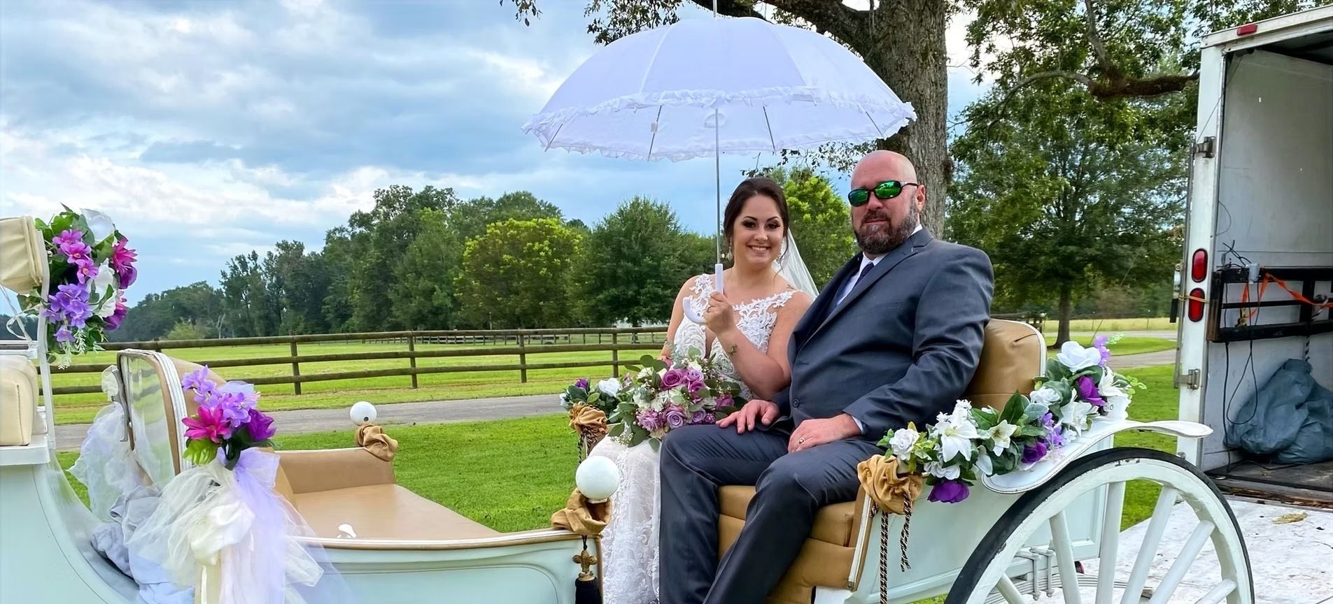 Couple seated in a decorated white carriage, sharing a lavender umbrella in a park-like setting.