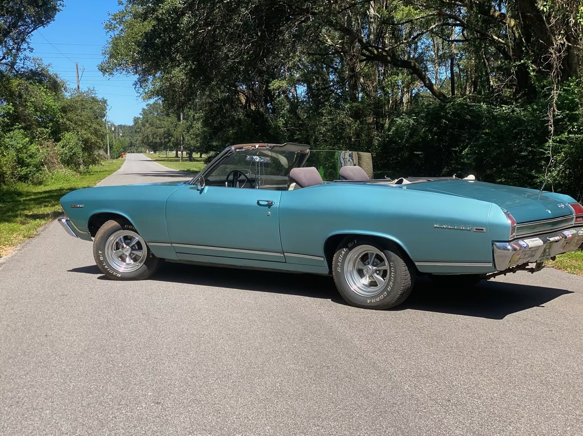 Turquoise classic convertible parked on a tree-lined road in sunlight