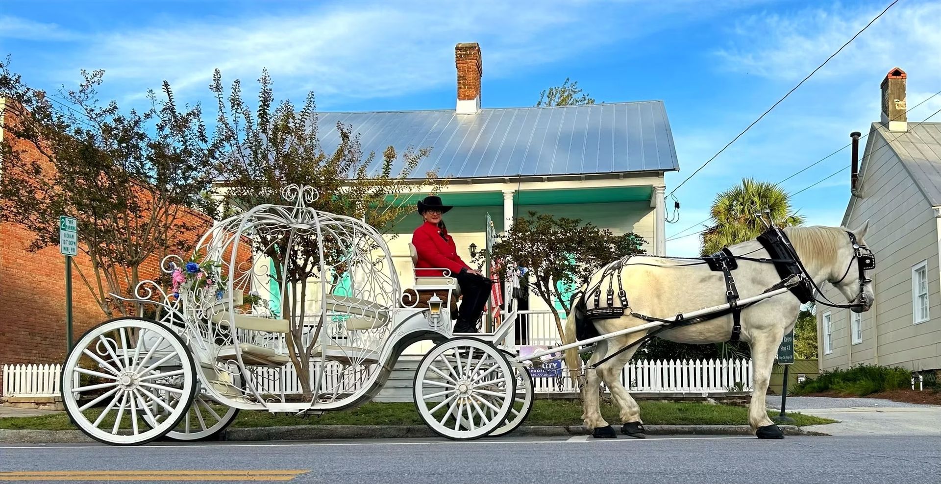 White horse pulls a white carriage with a driver past a house on a sunny street