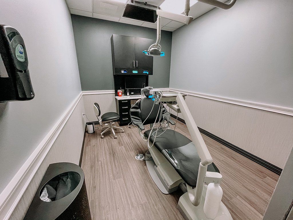 A dental office room with a chair, equipment, and cabinets. Gray walls, wood floor, and overhead light.