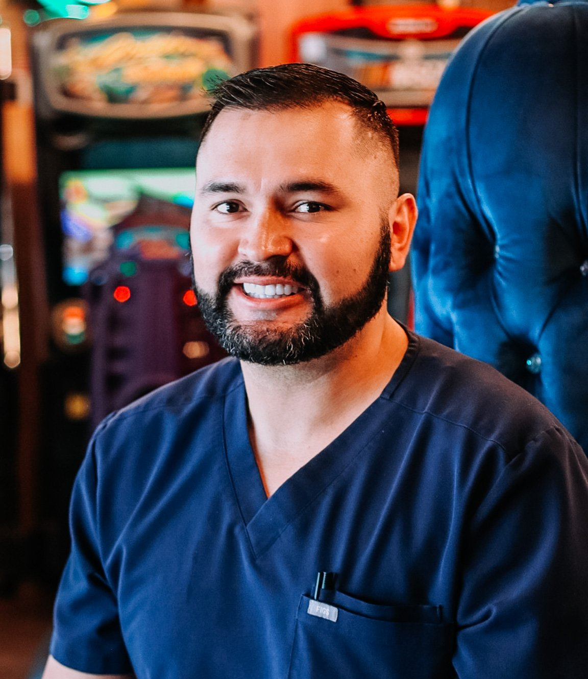 Man with dark hair and beard smiling, wearing blue scrubs, indoors near arcade games.