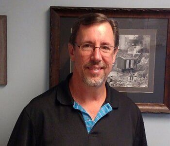 A man wearing glasses and a black shirt is standing in front of a framed picture.