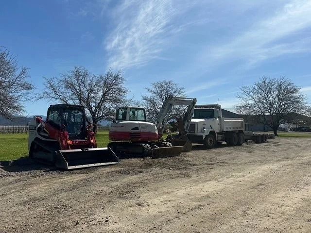 Red skid steer, beige machine, and white dump truck parked on a gravel lot under a blue sky.