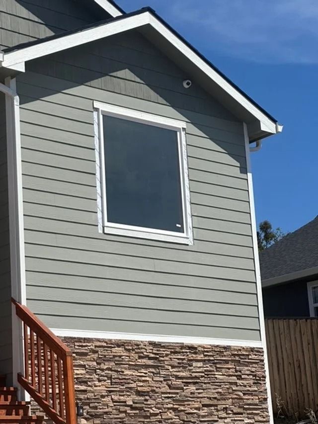 Gray house exterior with a white-trimmed window, stone lower facade, and blue sky.
