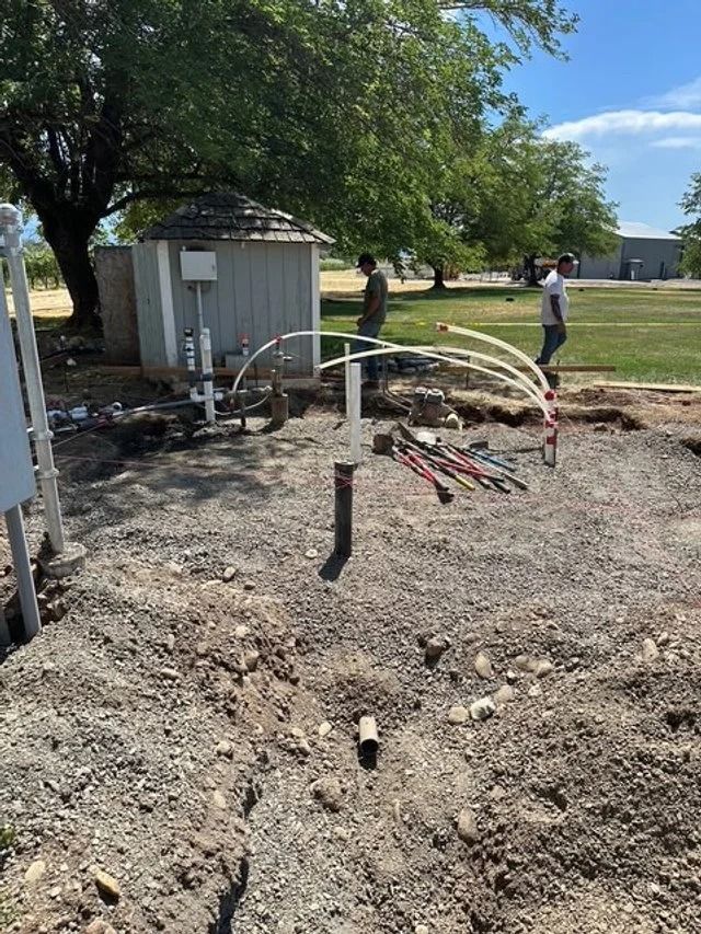 Construction site with a small shed, exposed pipes, and two workers on a dirt lot under trees.