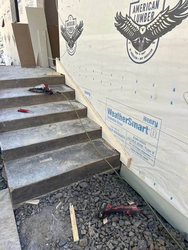 Partially finished stairs beside a white wall, with tools and gravel at the base.