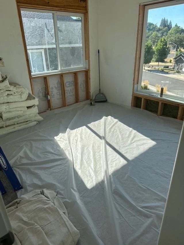 Empty room with plastic-covered floor, exposed wall studs, and sunlight through large windows
