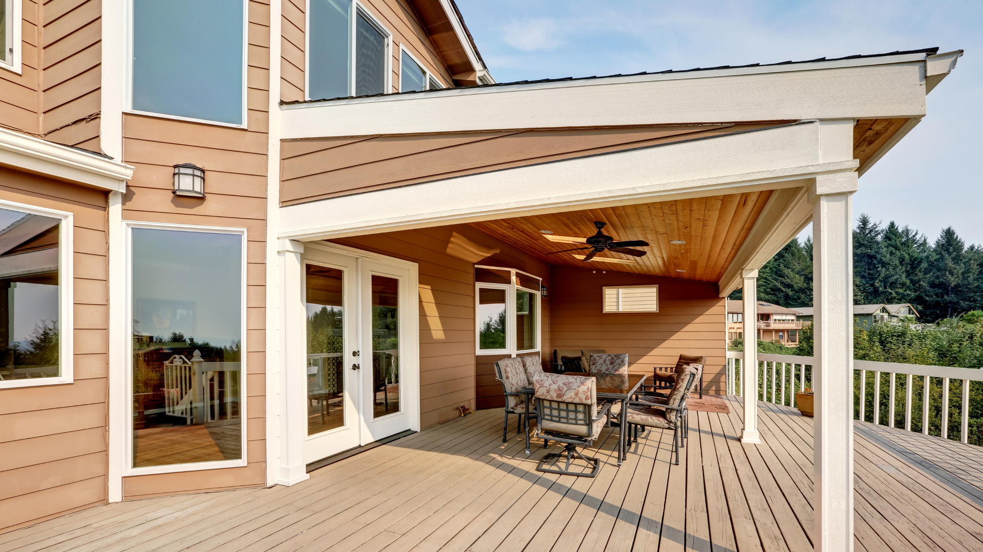 Wooden deck with a covered patio area, featuring a table, chairs, and a ceiling fan. Brown siding, white trim, and a forest in the background.
