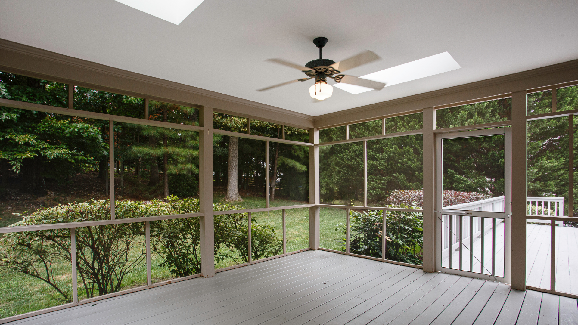 Screened porch with white ceiling fan, skylights, and green wooded view beyond railings