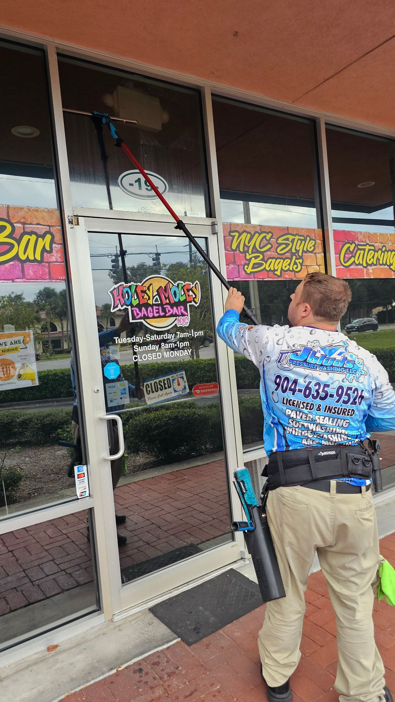 Man washes a storefront window with a long-handled brush. He wears a blue shirt, tan pants, and a tool belt.