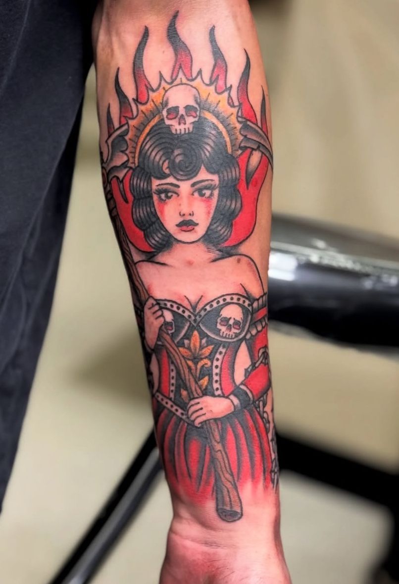 Forearm tattoo of a stylized woman with a flaming headdress, red accents, and ornate corset dress