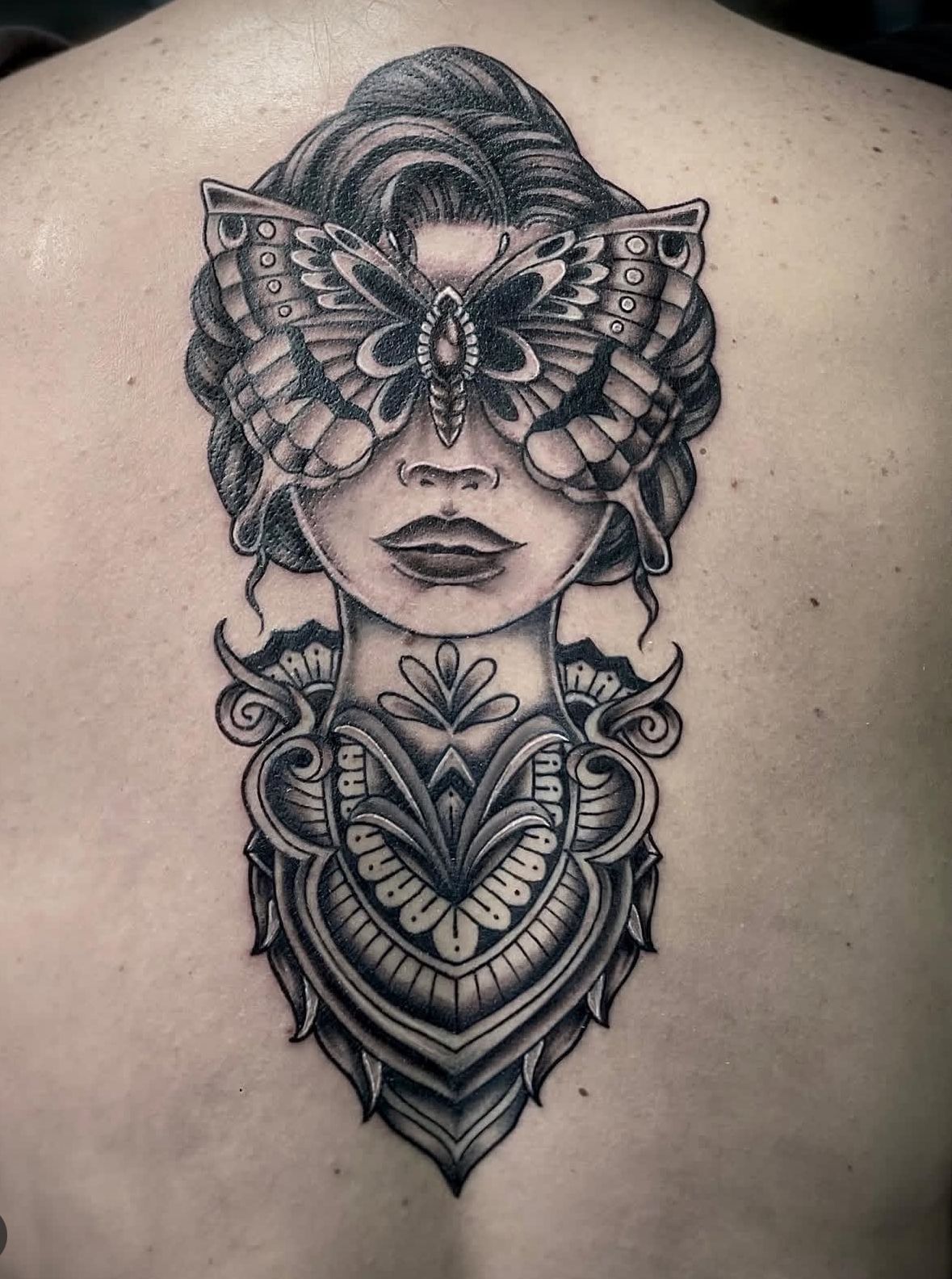 Black-and-gray back tattoo of a masked female face with ornate butterfly and floral patterns