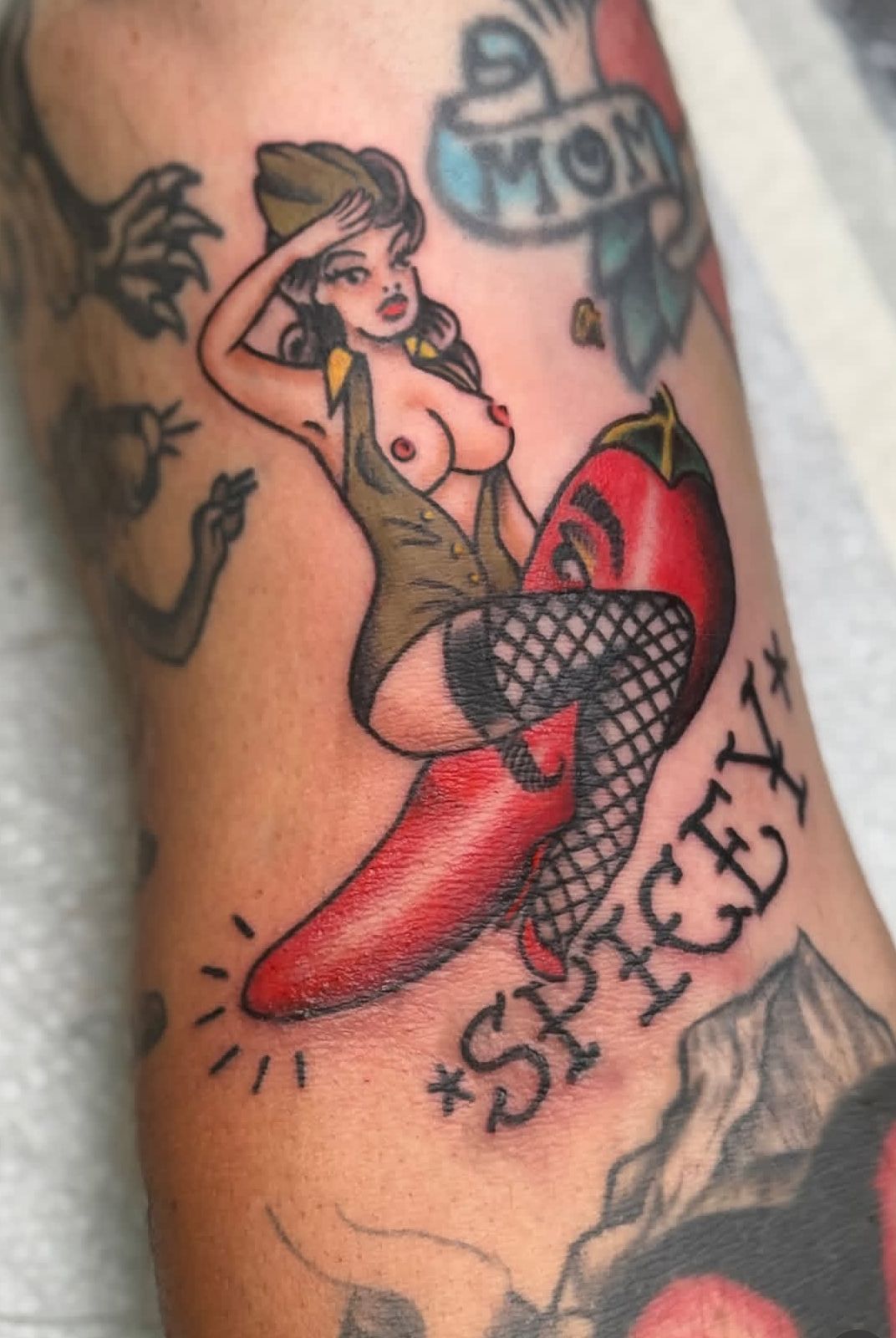 Pin-up style tattoo of a woman sitting on a red heart, with surrounding tattooed designs and script