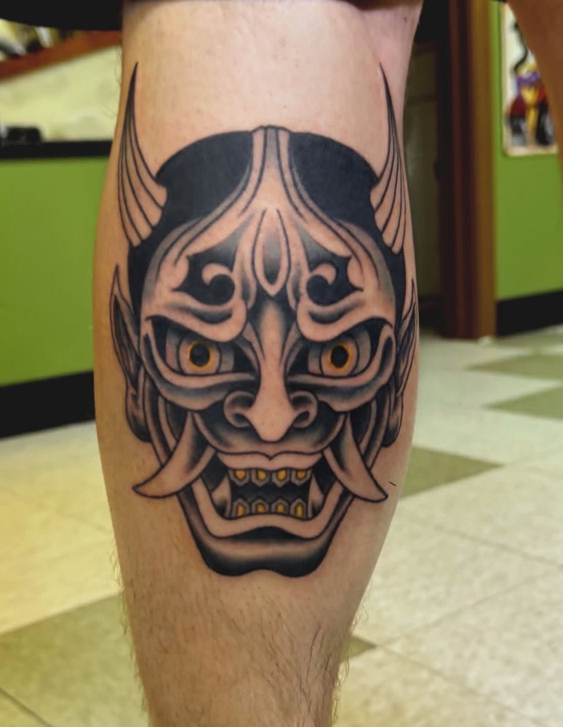 Black tribal demon face tattoo on a person’s calf, with sharp eyes and horns.
