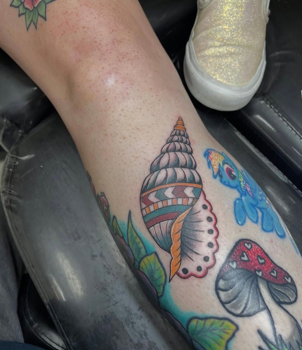 Leg with colorful sea-themed tattoos, including a shell and mushroom, resting on a black chair.