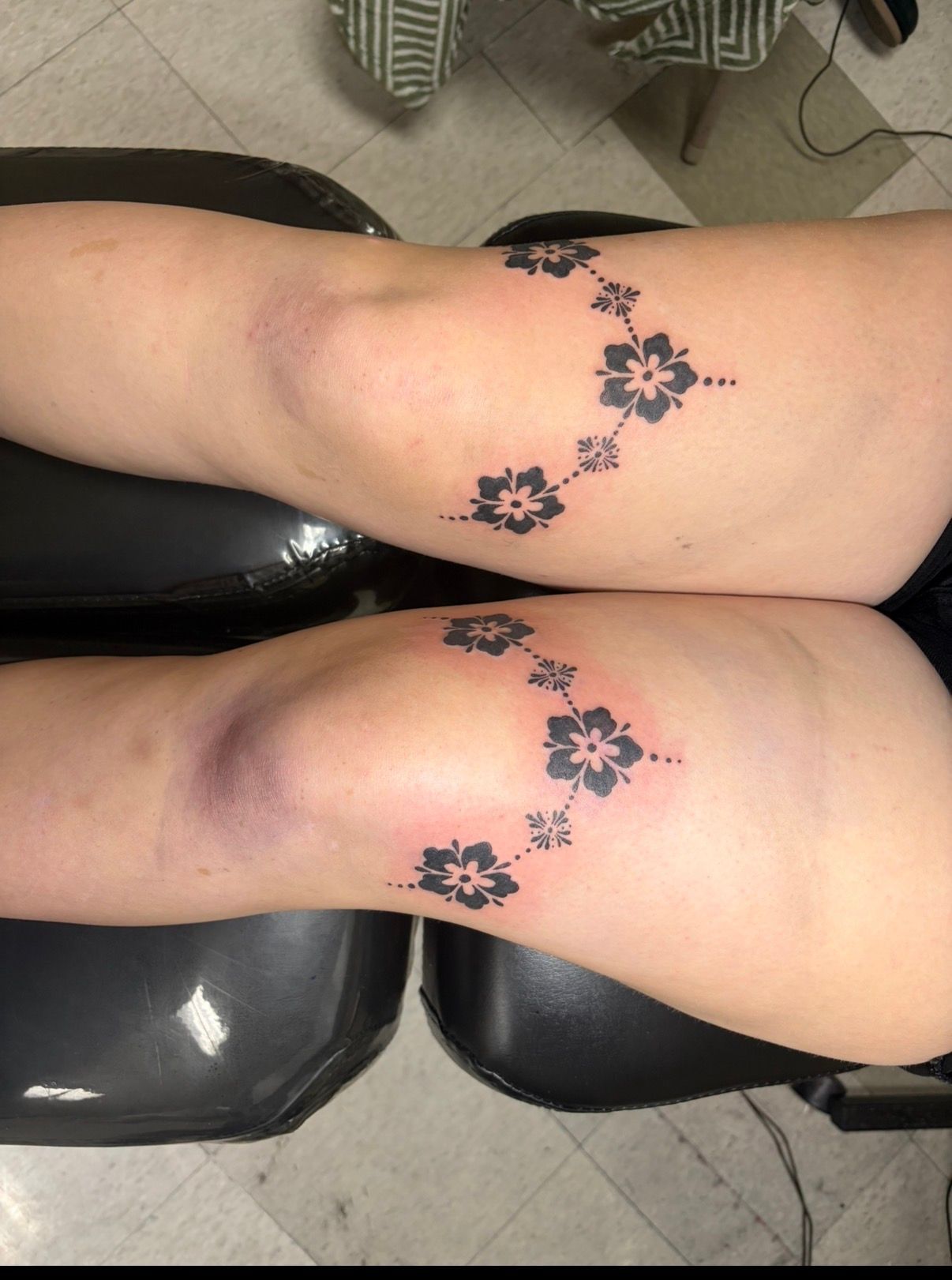 Close-up of two tattooed legs with dark floral designs on the thighs, resting on black chairs.