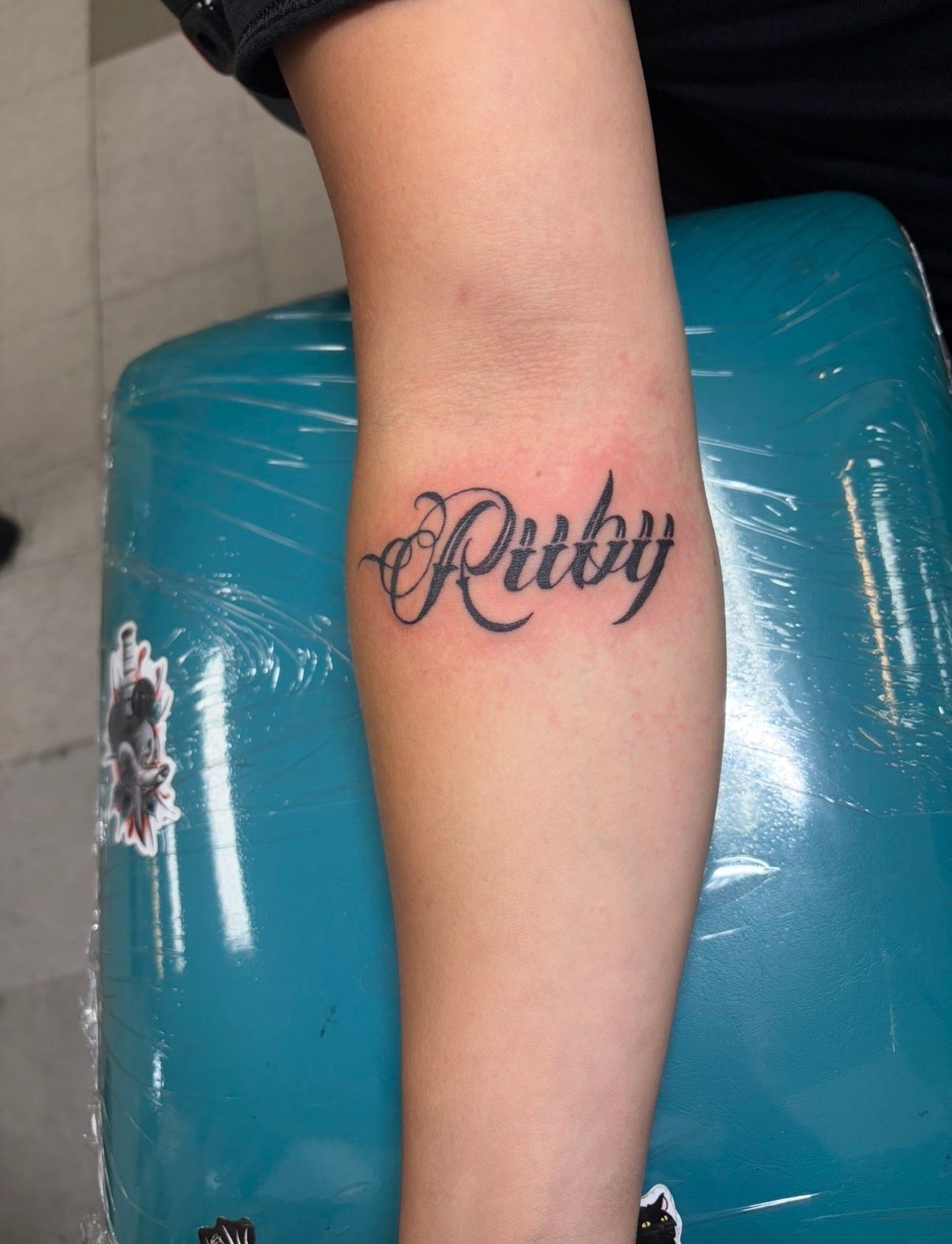 Forearm tattoo reading “Pitby” in black script on skin against a teal chair.