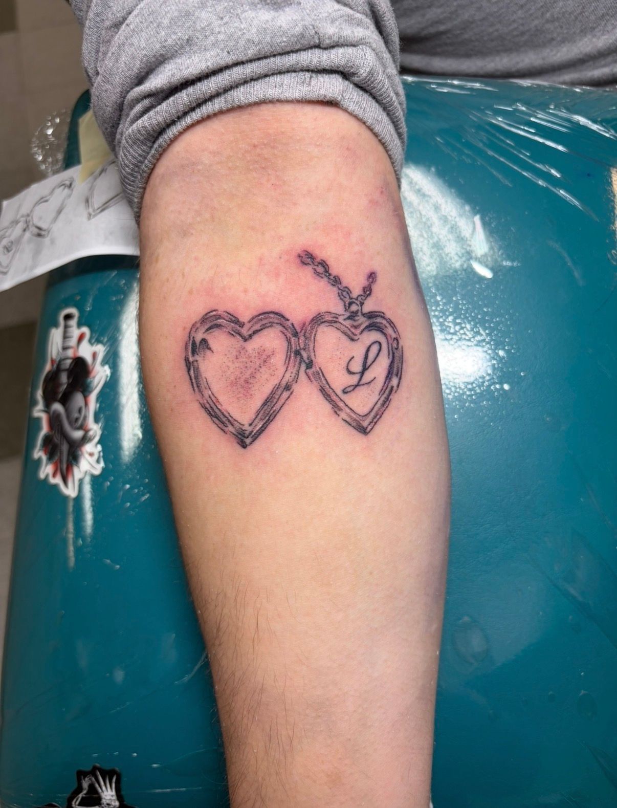 Forearm with two heart-shaped tattoos, one with a treble clef, resting against a teal chair.