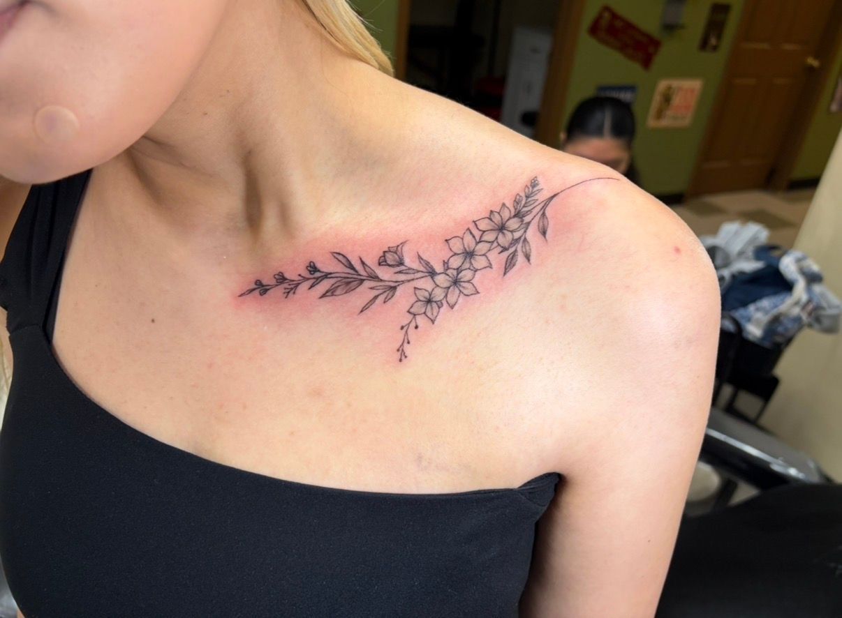 Shoulder tattoo of a leafy vine in black ink on a person wearing a black off-shoulder top
