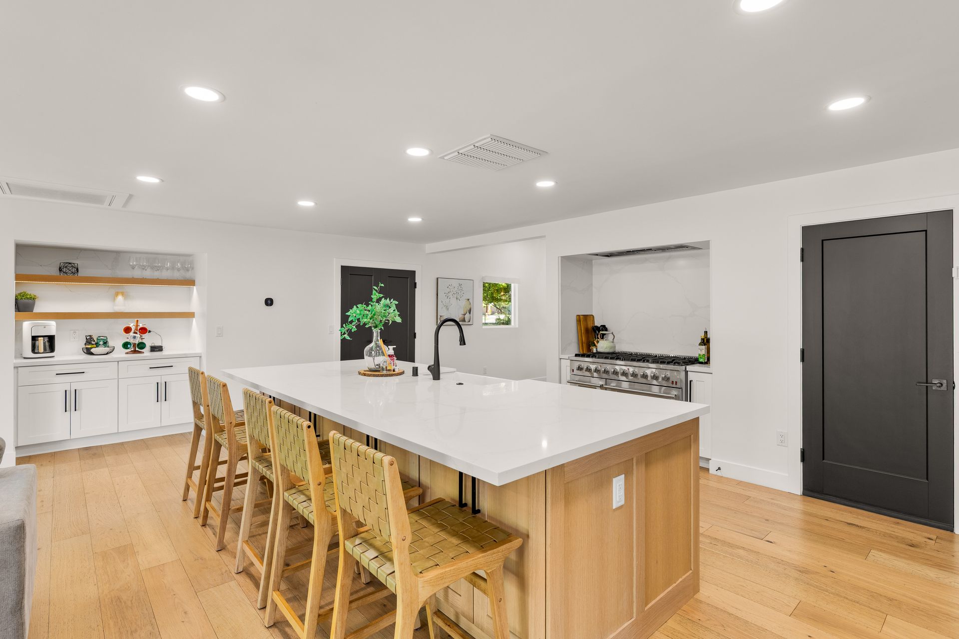 Bright modern kitchen with white island, wooden stools, light wood floors, and dark pantry door
