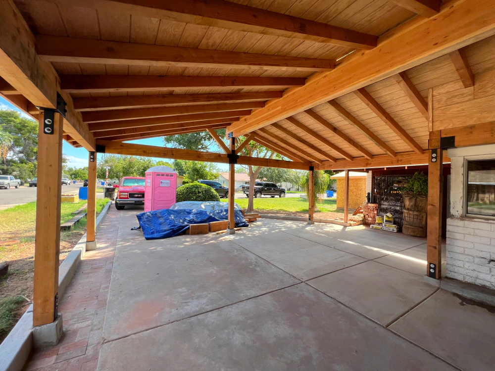 Covered patio with wood beams, concrete floor, and a pink playhouse beside a blue tarp outside.
