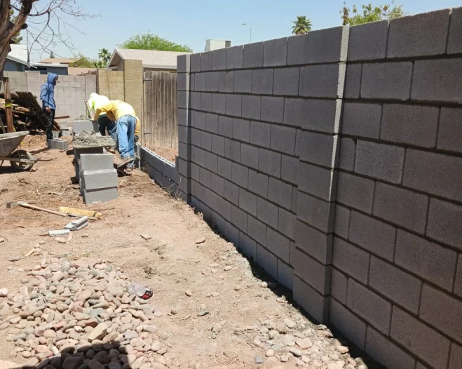 Workers stacking gray concrete blocks along a construction site wall beside a dirt path