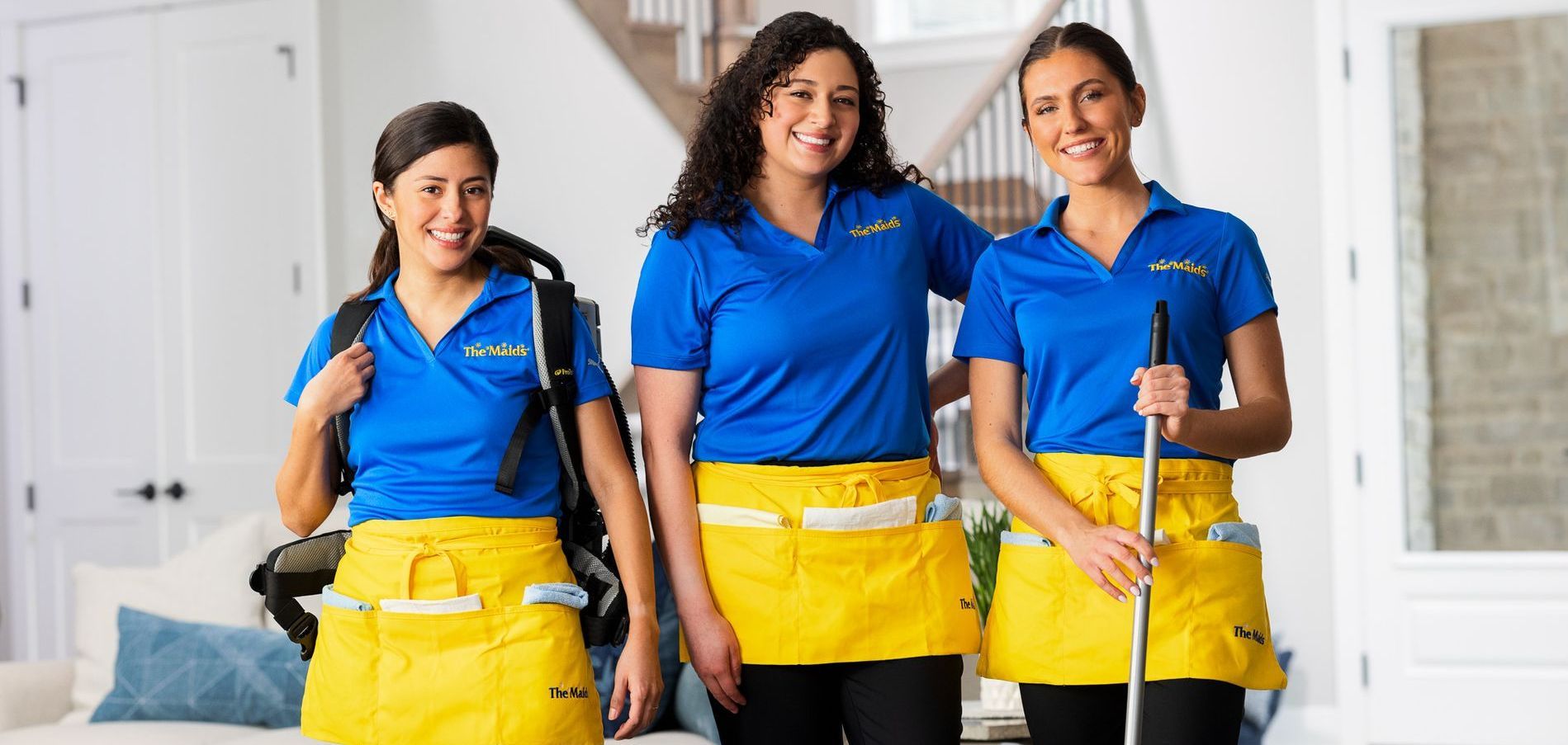 Three house cleaners in blue shirts and yellow aprons standing in a living room, smiling and holding cleaning tools