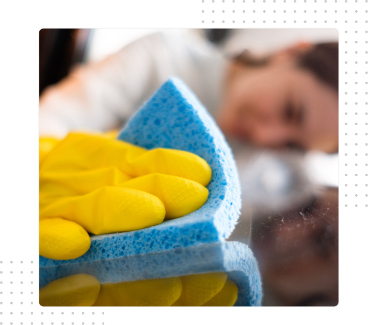 Yellow-gloved hand wiping a window with a blue sponge, blurred person in the background