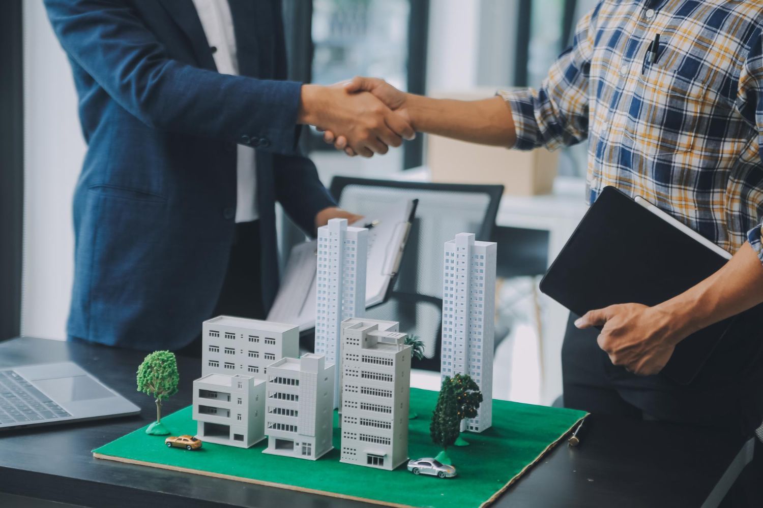 Two professionals shake hands over a scale model of a city development on a desk in an office setting.