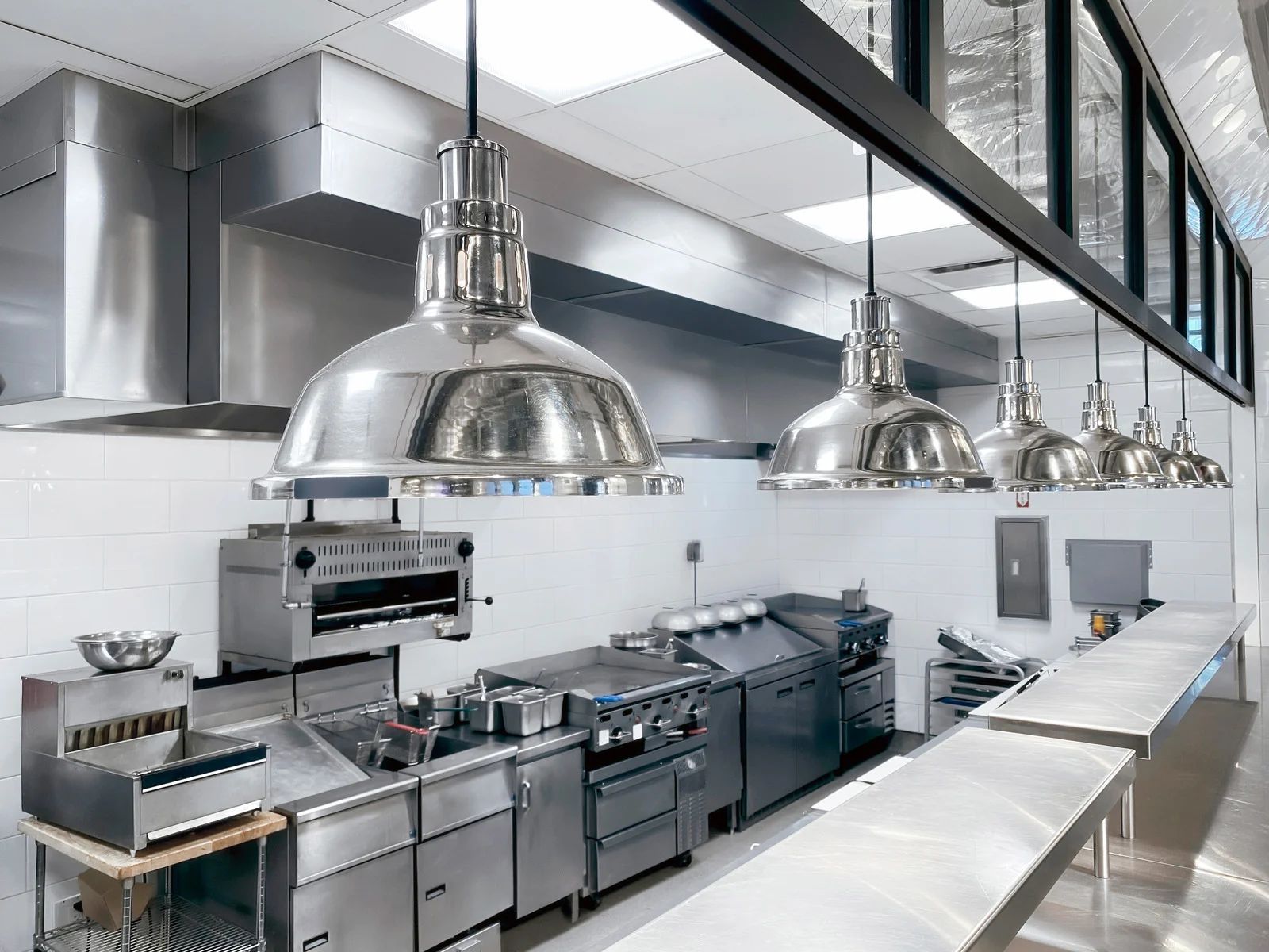 Modern stainless-steel commercial kitchen with large hanging heat lamps and prep counters