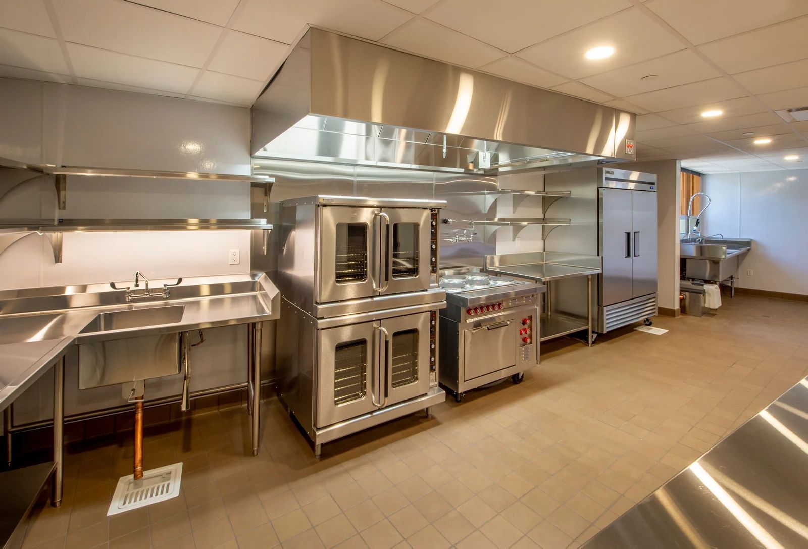 Commercial kitchen with stainless steel counters, ovens, sinks, and a prep area under bright ceiling lights