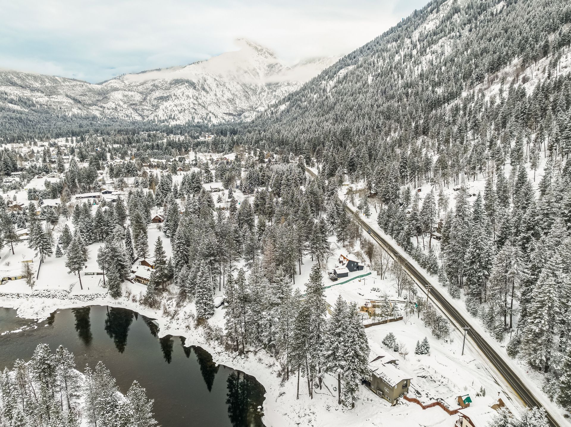 Snow-covered mountain valley with a river, road, and trees; overcast sky.