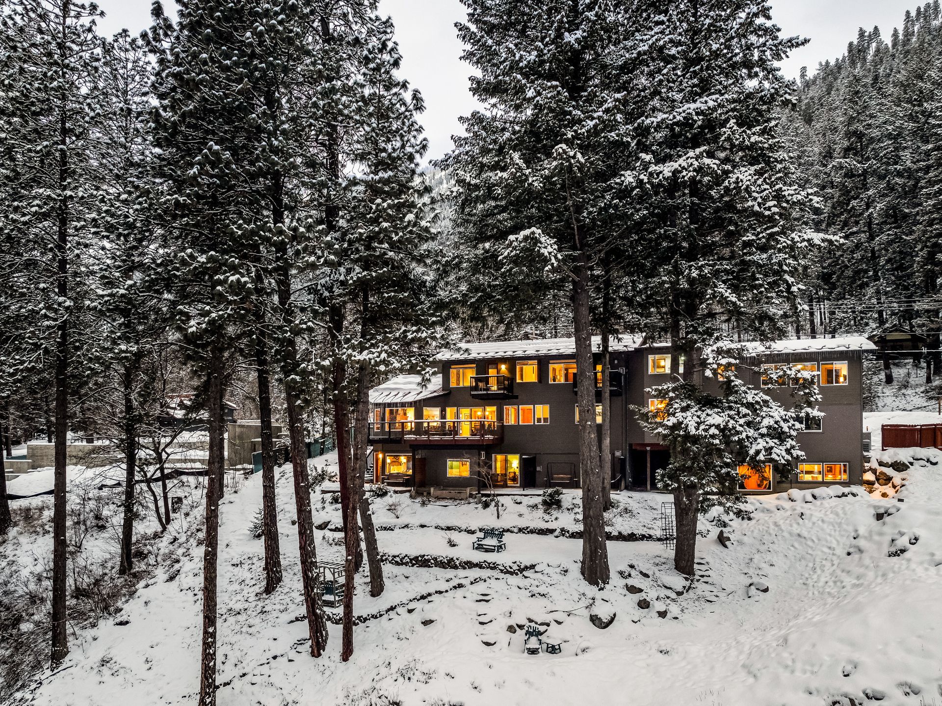 Snow-covered hotel nestled among tall pine trees on a hillside; lights glowing inside, winter setting.