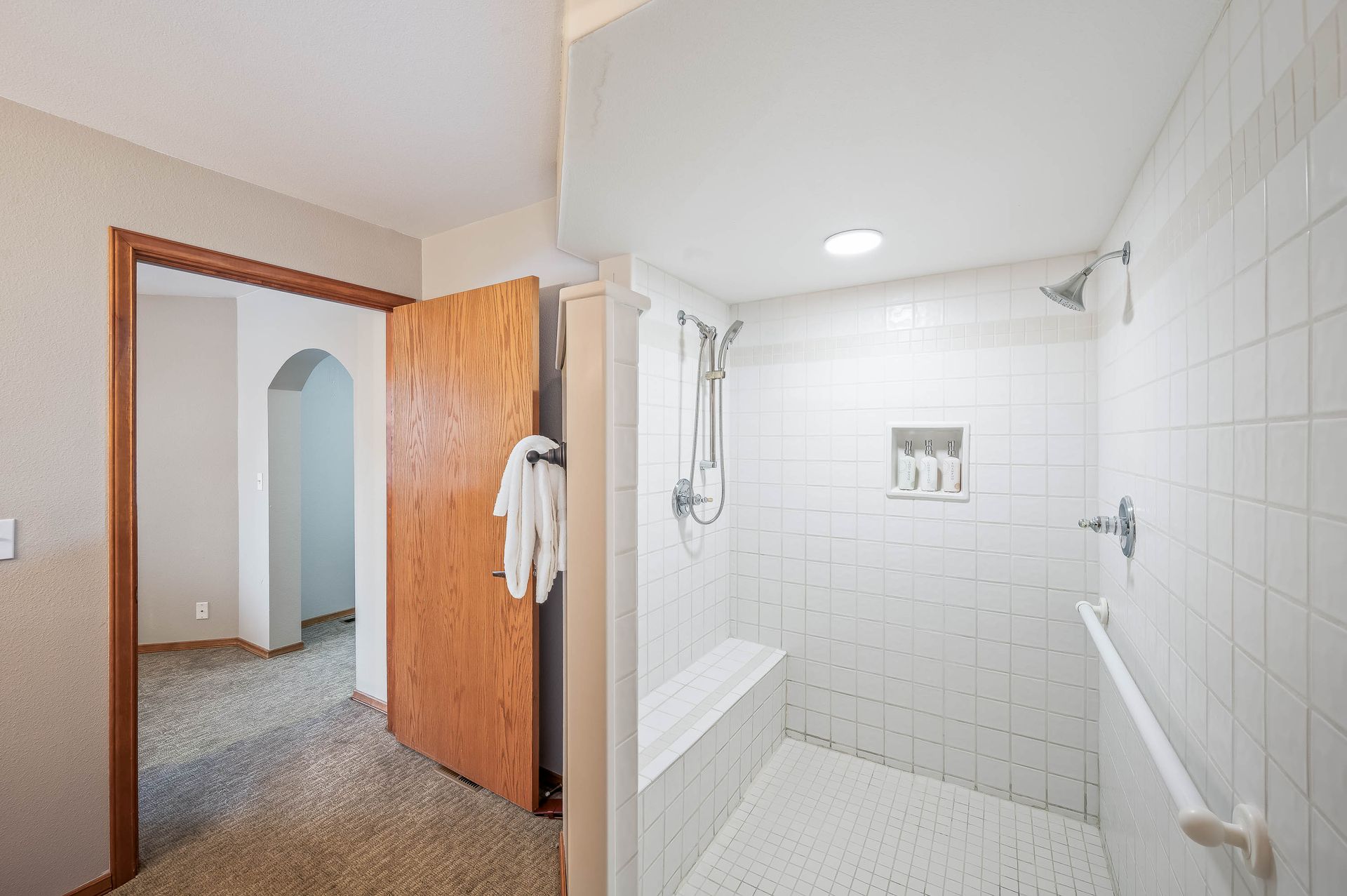 Bathroom interior with a white-tiled shower, white bench, and open door to hallway with brown door.