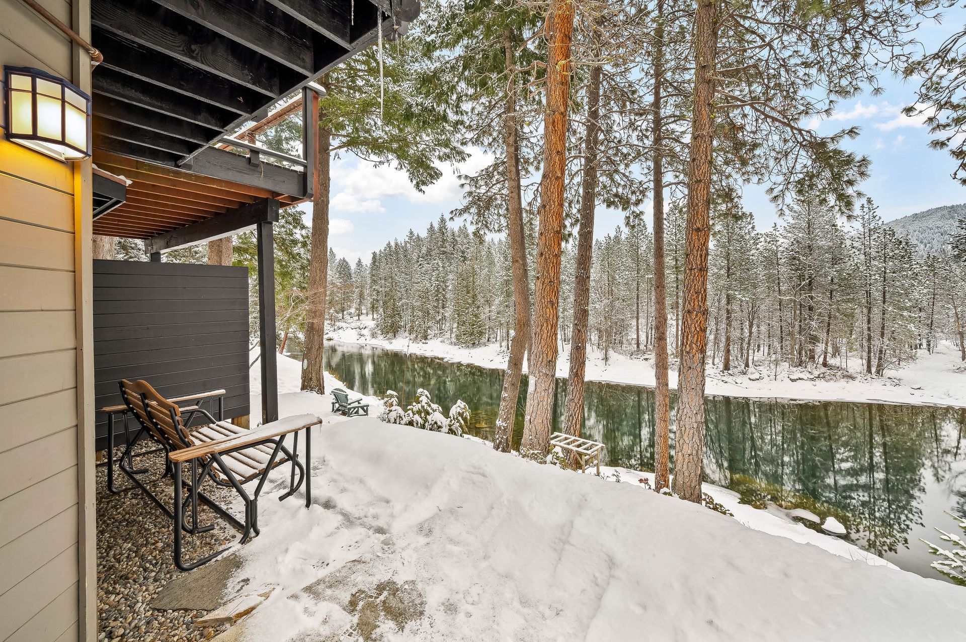 Snowy patio overlooking a river lined with trees. Two chairs and a small table on the patio.