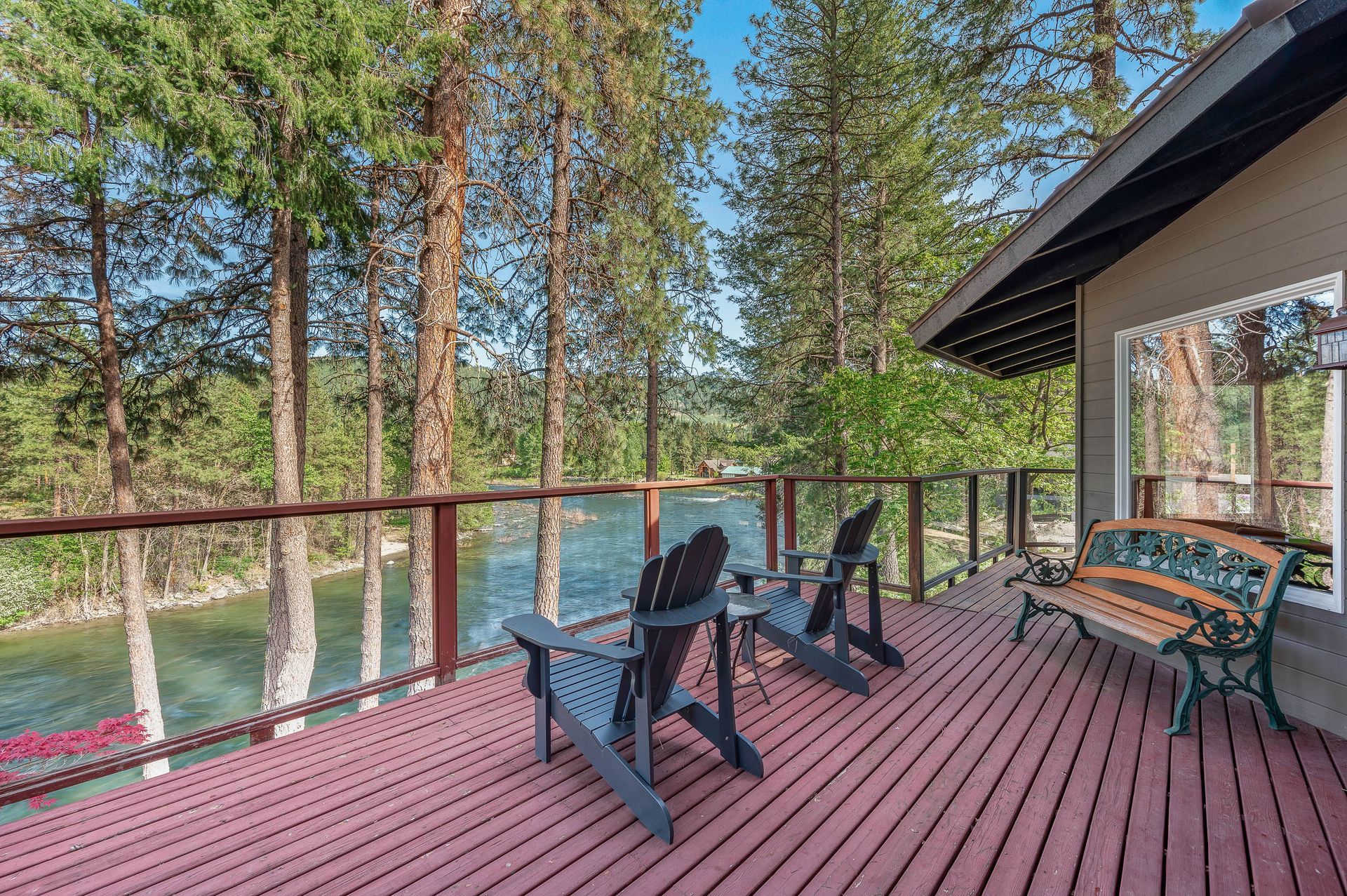 Wooden deck with black chairs and green bench overlooking a forest and water through a railing