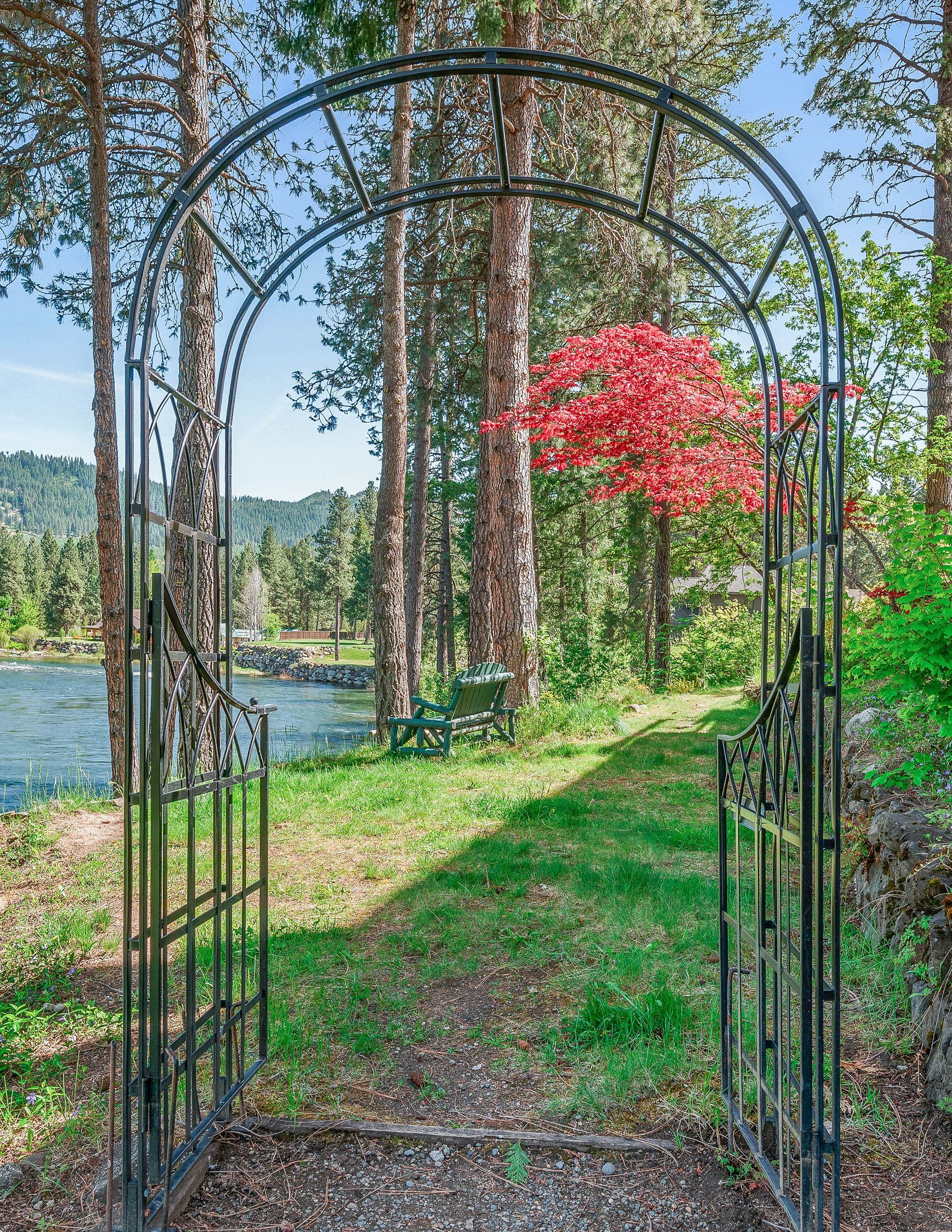 Iron gate archway leading to a path beside a river. Red tree visible. Green grass, trees, and blue sky.