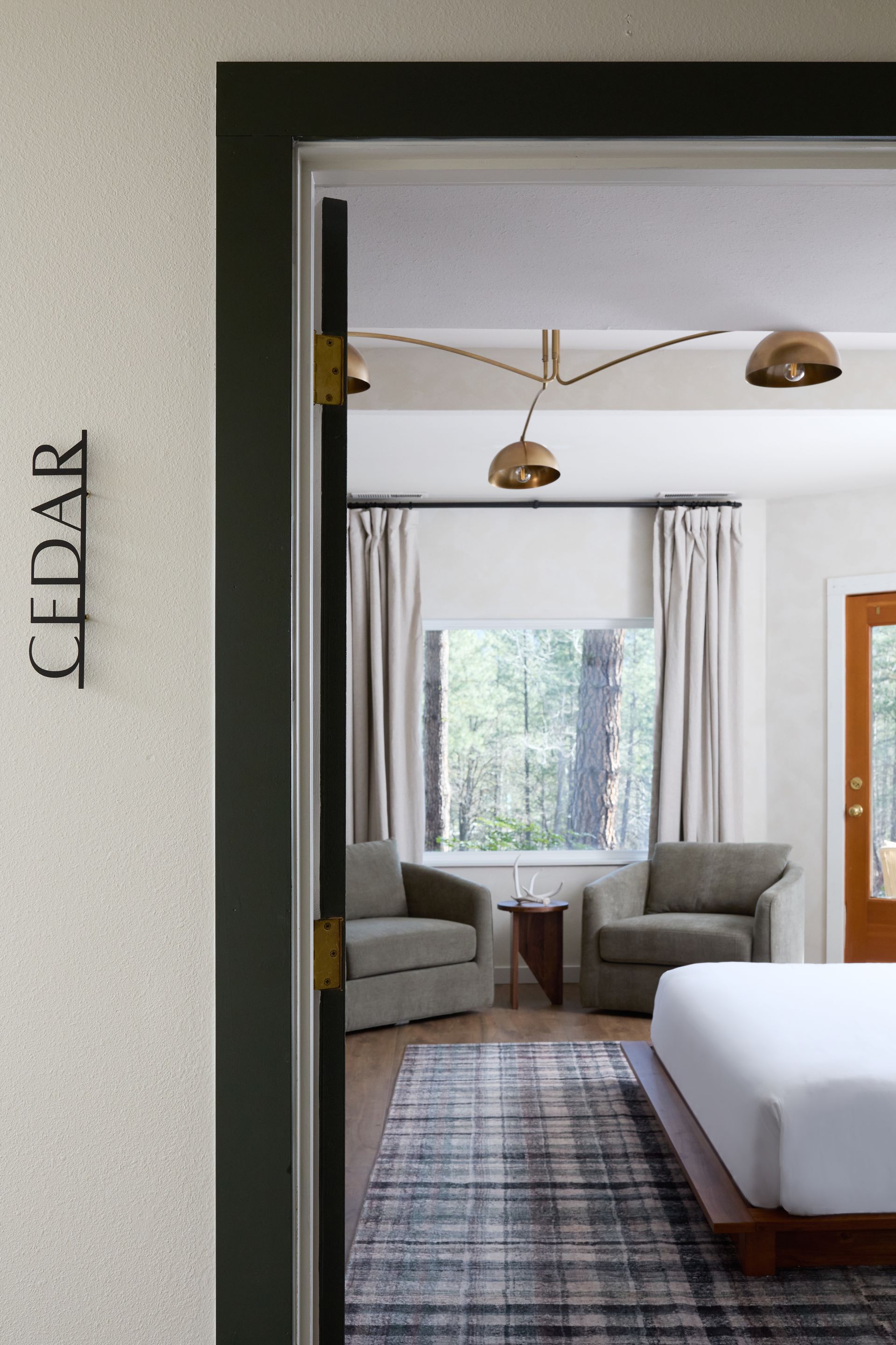 Modern bedroom viewed through a doorway, with gray chairs, a window, and brass pendant lights.