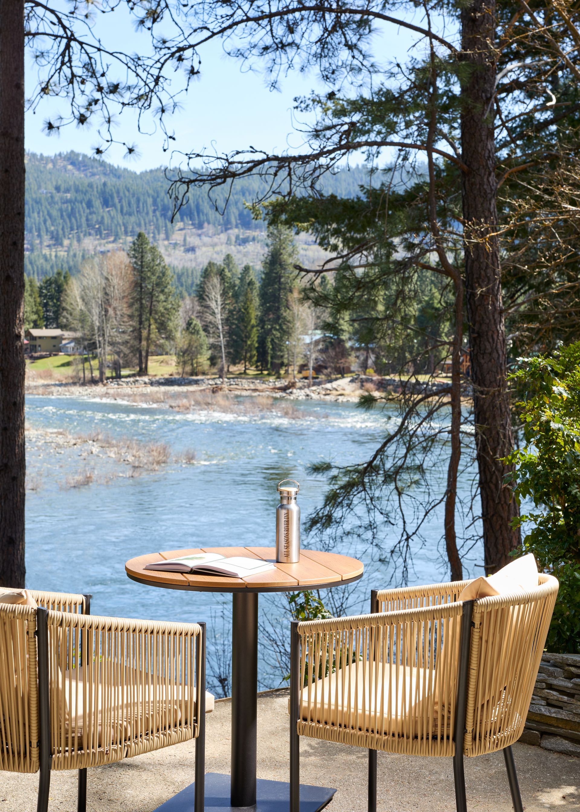 Riverside patio with wicker chairs and a small table overlooking a blue river and pine-covered hills