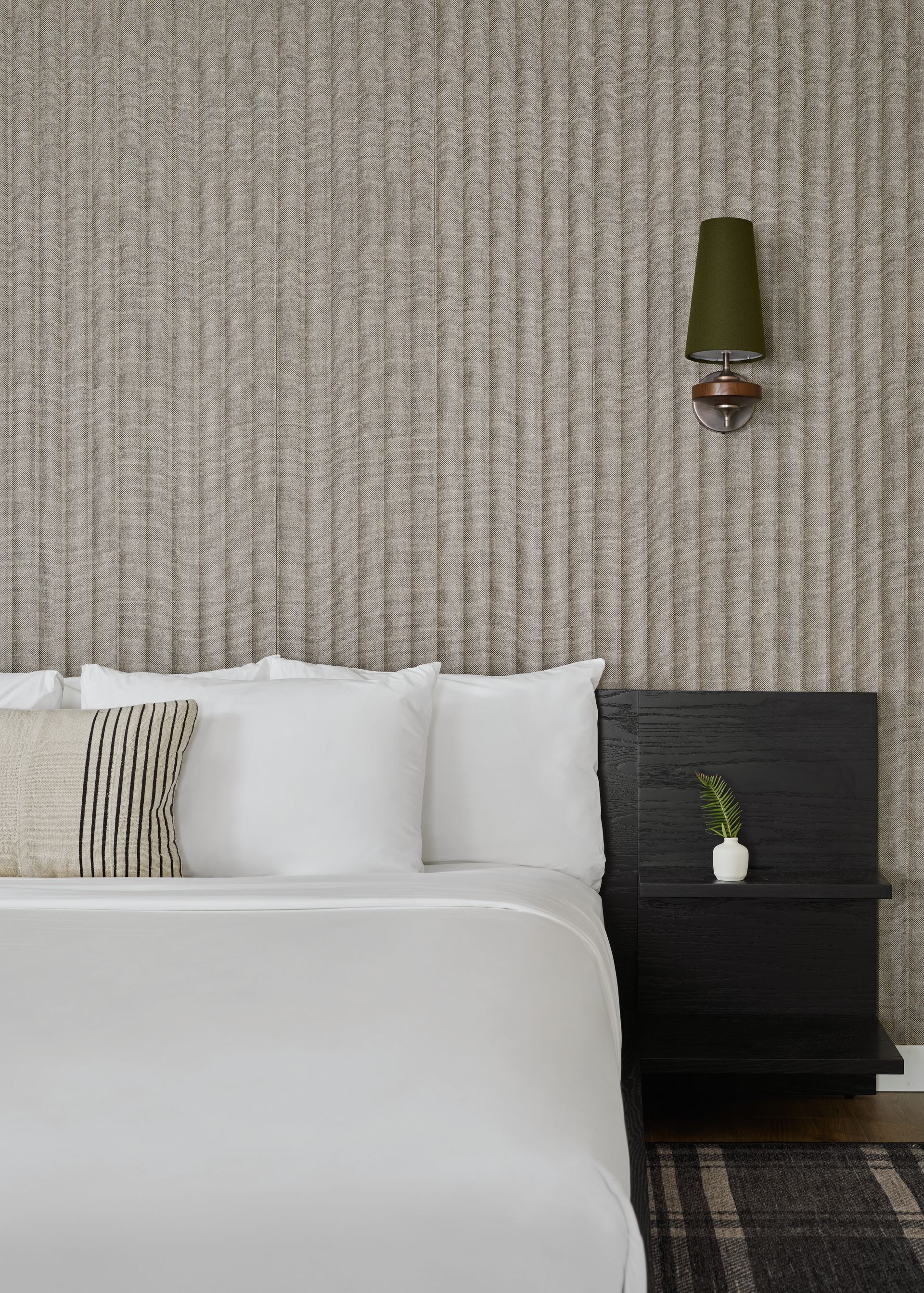 Modern bedroom with white bedding, textured wall, and a wall lamp above a bedside table.