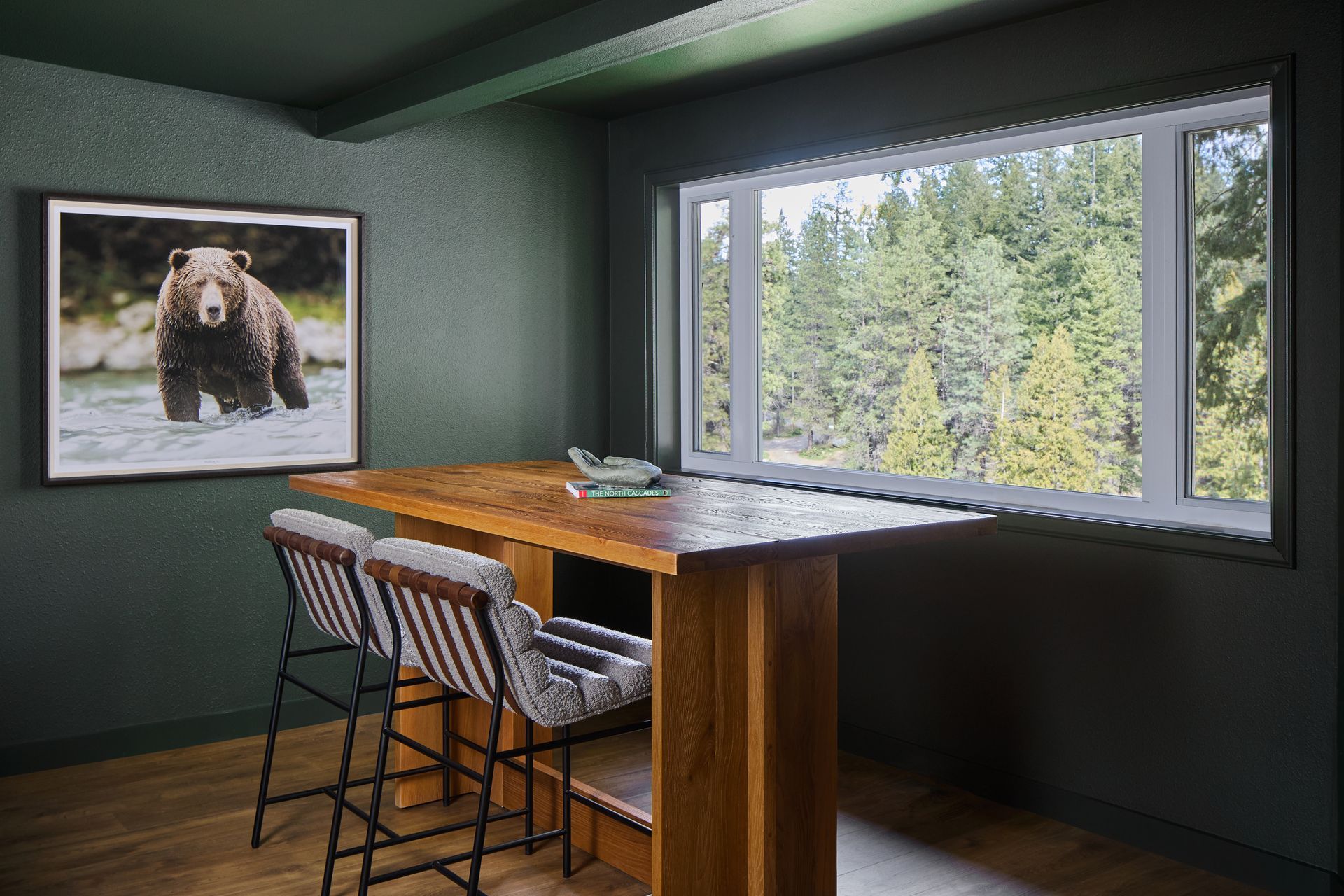 Cozy room with wooden counter stools, bear wall art, and a large window overlooking evergreen trees.