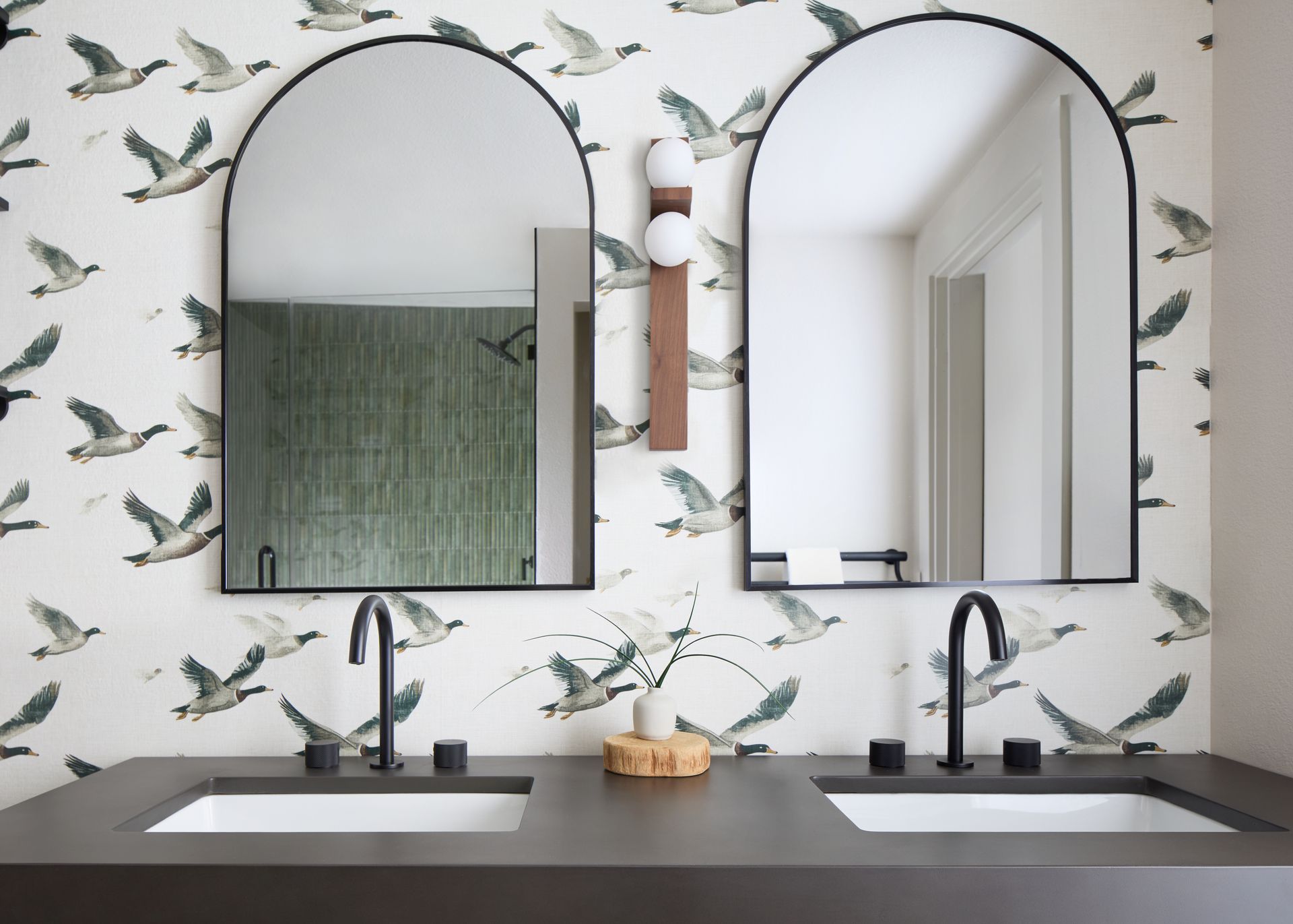 Modern bathroom vanity with twin arched mirrors, black faucets, and botanical wallpaper.