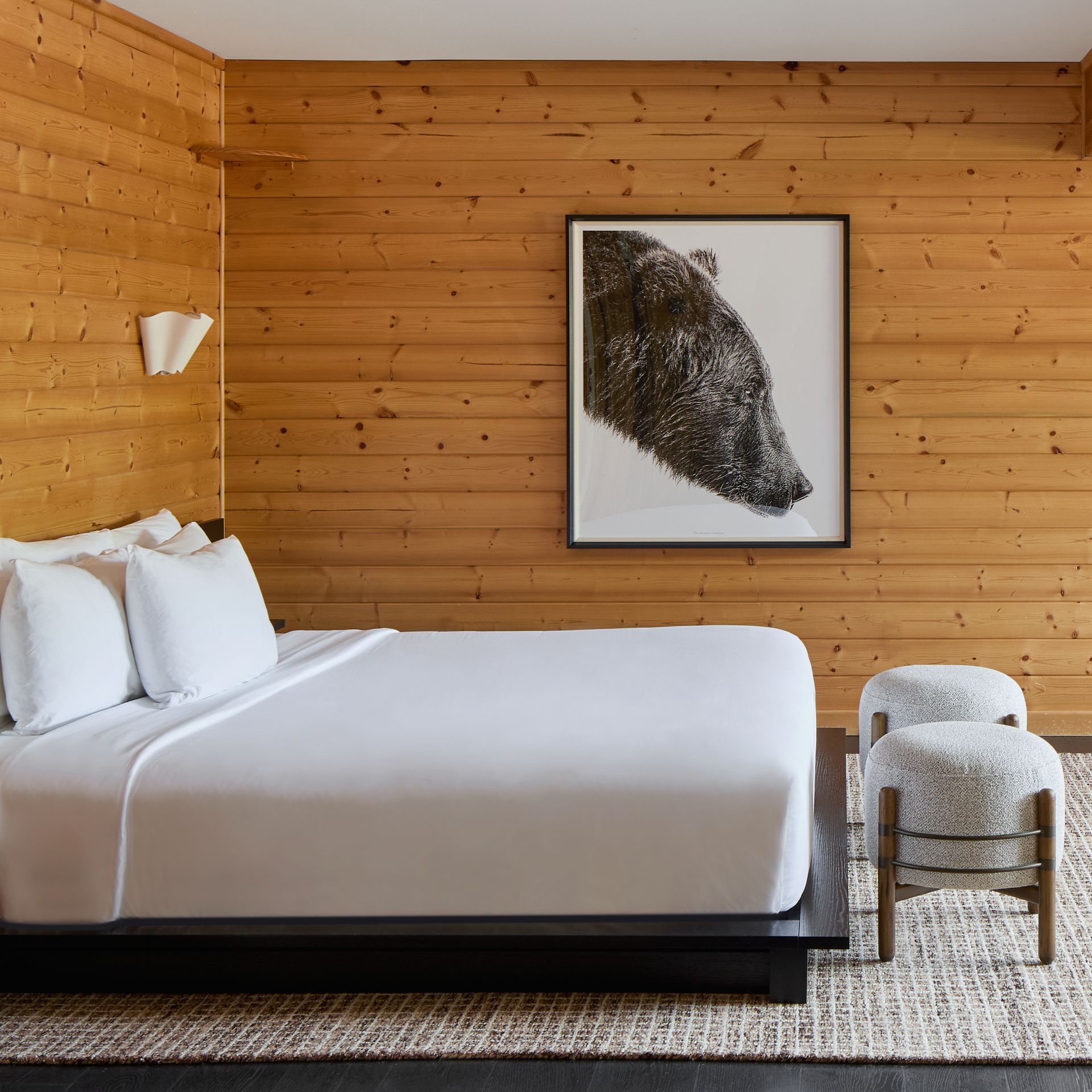 Minimalist bedroom with wood-paneled walls, white bed, framed art, and ottomans on a textured rug
