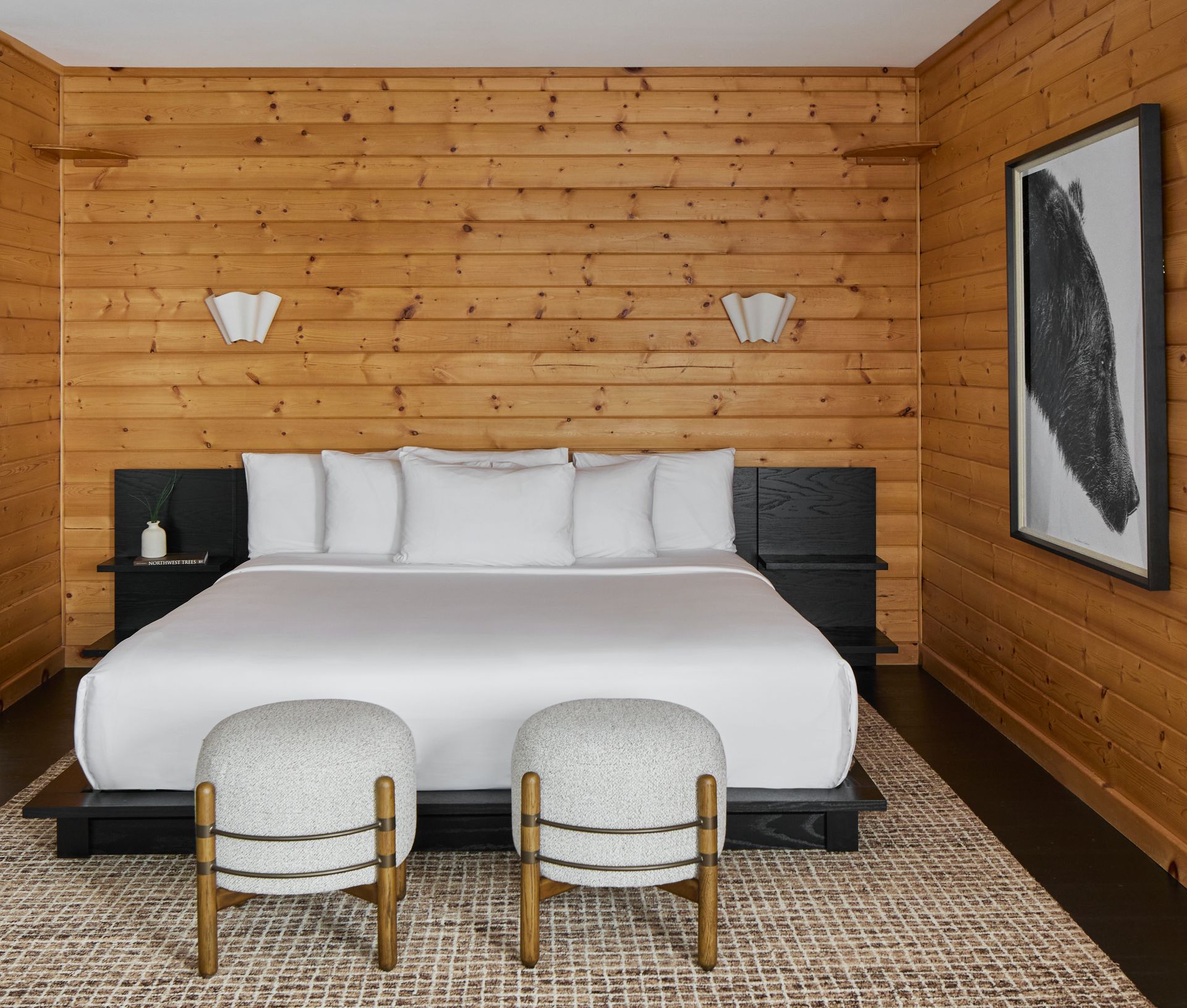 Modern bedroom with wooden walls, white bed, black nightstands, framed art, and two upholstered stools.
